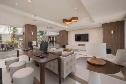 Sala de estar moderna e iluminada com sofás brancos, detalhes em madeira, janelas amplas e TV fixada na parede.