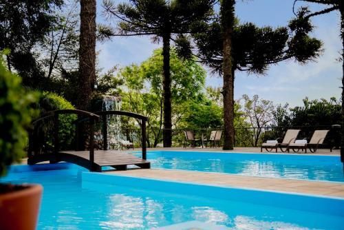 Piscina externa ensolarada com água azul, uma pequena ponte, espreguiçadeiras e árvores ao fundo.