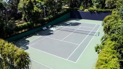 Quadra de tênis externa vazia, cercada por árvores, com superfície de jogo verde e cinza.