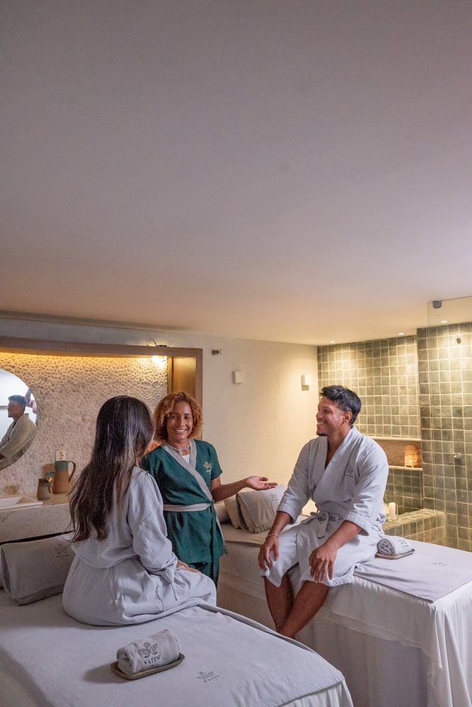 Três pessoas conversando em uma sala de tratamento de spa, sentadas em macas de massagem com toalhas e iluminação suave.