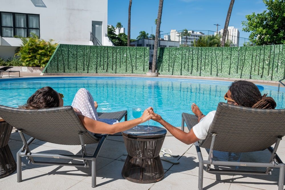 Duas pessoas relaxando em espreguiçadeiras à beira de uma piscina azul, de mãos dadas sobre uma pequena mesa.