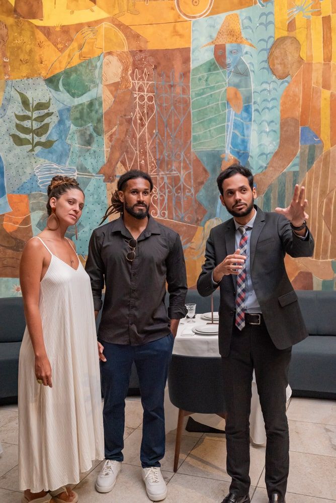 Três pessoas em pé em frente a um mural colorido em um evento em local fechado, uma delas segurando um microfone.