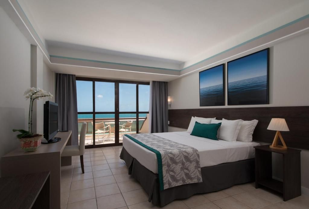 Quarto de hotel com cama king-size, varanda com vista para o mar, escrivaninha e decoração de parede azul.