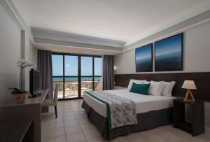 Quarto de hotel moderno com cama king-size, vista da varanda, paredes cinza, detalhes em azul-petróleo e o oceano ao longe.