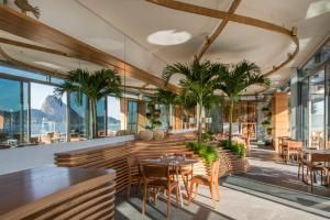 Café moderno e luminoso com mesas de madeira, palmeiras em vasos e grandes janelas com vista para as montanhas e o mar.