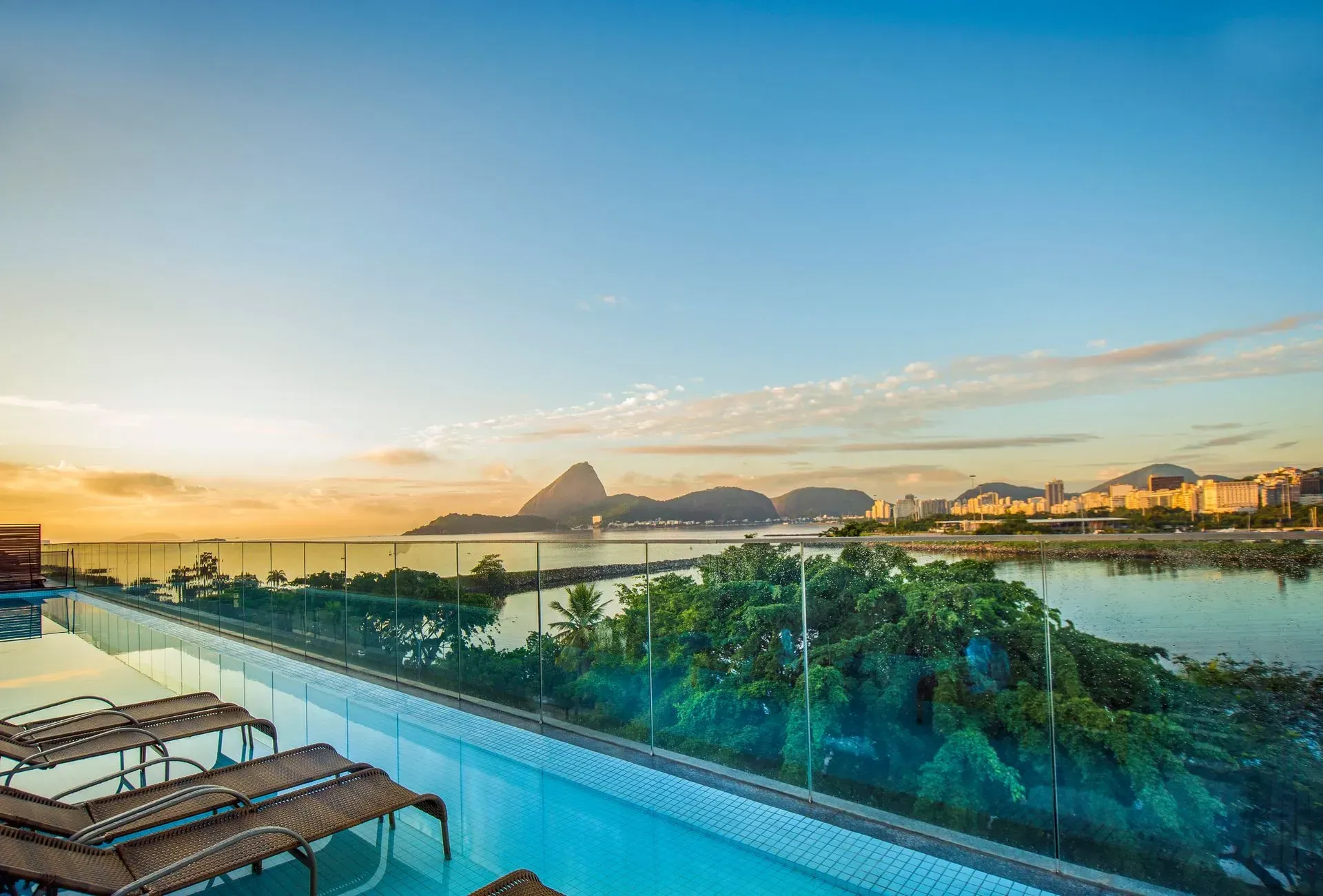 Piscina infinita com vista para a baía tropical ao pôr do sol, com palmeiras e montanhas ao longe.