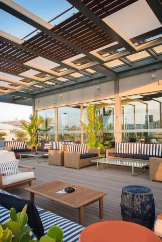Lounge moderno na cobertura com sofás listrados, mesas de madeira, plantas e um pergolado ao pôr do sol.