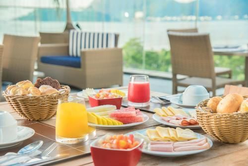 Café da manhã completo com suco, pão, frutas e doces em uma mesa perto de uma janela ensolarada para o pátio.