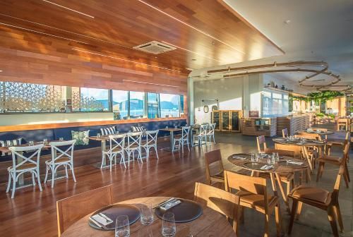Interior de café moderno com teto de madeira, mesas brancas e grandes janelas com vista para uma cidade iluminada.