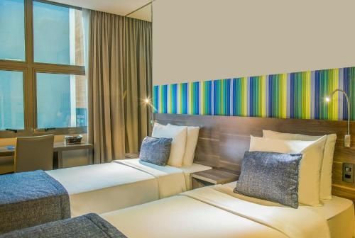 Quarto de hotel moderno com duas camas de solteiro, detalhes em azul e uma grande janela com vista para a cidade.