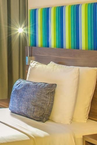 Cama de hotel com travesseiros brancos, almofada decorativa azul e aplique de parede com cabeceira listrada e colorida.