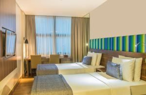 Quarto de hotel moderno com duas camas, cabeceira listrada colorida, assento junto à janela e decoração em tons suaves de bege.
