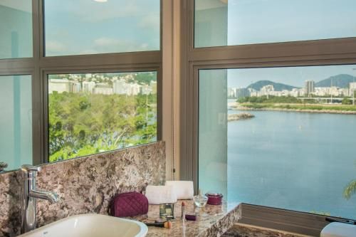 Vista da janela do banheiro para um lago tranquilo e colinas ao longe, com uma pia de granito e artigos de higiene pessoal em primeiro plano.