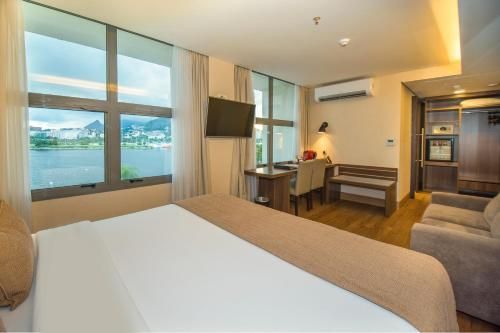 Quarto de hotel com amplas janelas com vista para o lago, cama, escrivaninha, TV e área de estar.