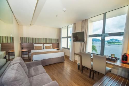 Quarto de hotel moderno com cama, sofá cinza, mesa de jantar e grandes janelas com vista para o mar.