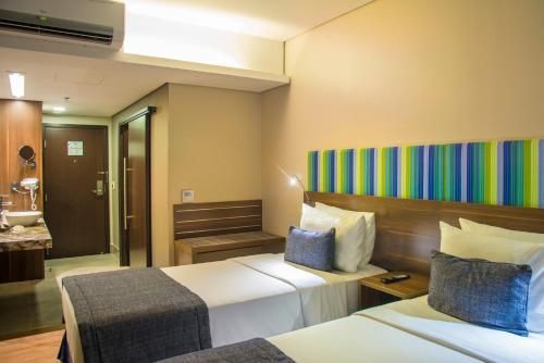 Dois quartos de hotel bege com duas camas de solteiro, cabeceira listrada e cômoda perto da porta.