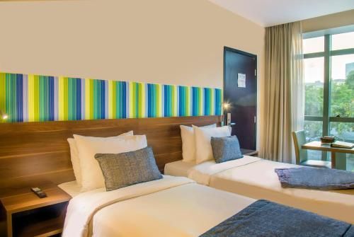 Quarto de hotel luminoso com duas camas de solteiro, cabeceira listrada e janela grande com cortinas.