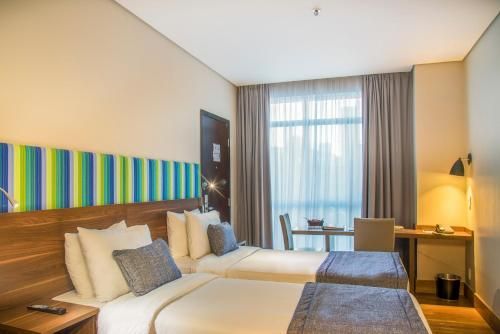 Quarto de hotel moderno com duas camas de solteiro, cabeceira colorida, luminária de mesa e janela grande com cortinas.