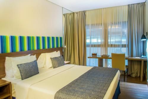 Quarto de hotel moderno com cama king-size, cabeceira listrada, escrivaninha junto à janela e cortinas bege.