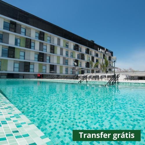 Piscina de hotel com água turquesa ao lado de um prédio moderno sob um céu azul claro; placa “Transfer grátis”