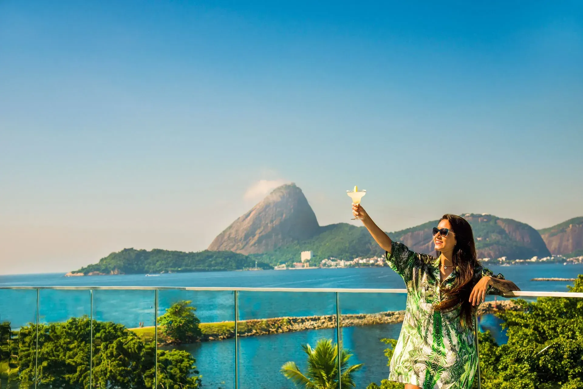 Mulher posando com o braço erguido em uma baía tropical com montanha sob um céu azul.