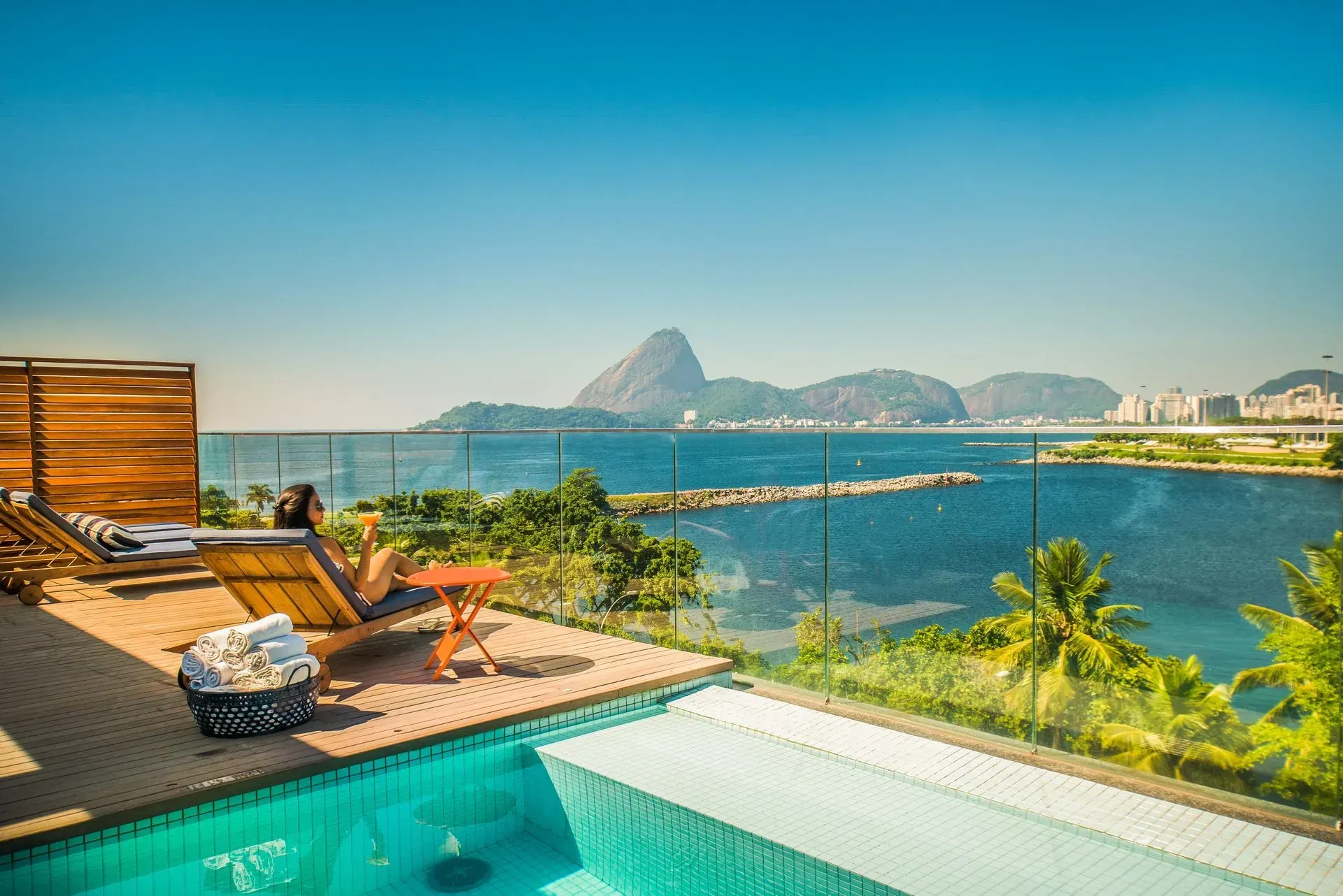Terraço de piscina luxuoso com vista para o oceano e montanhas ao longe ao pôr do sol.