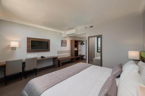 Quarto de hotel moderno com cama, escrivaninha, cadeiras, TV e janela grande.