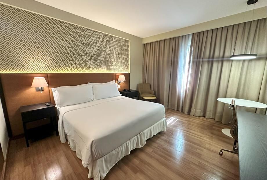 Quarto de hotel moderno com cama branca, abajures, cortinas e uma pequena escrivaninha junto à janela.