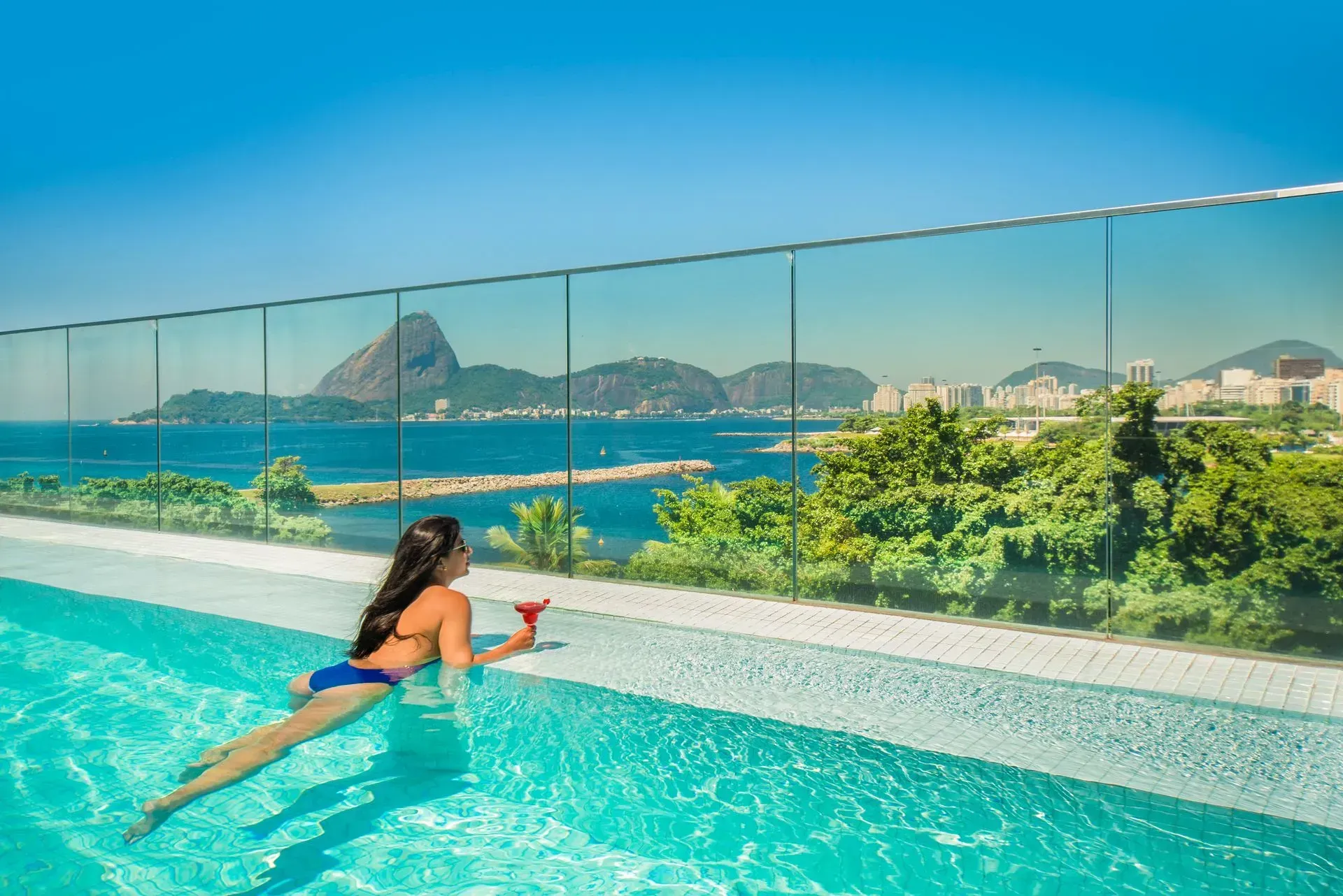 Mulher relaxando em uma piscina infinita com vista para o oceano, montanhas e uma cidade litorânea.