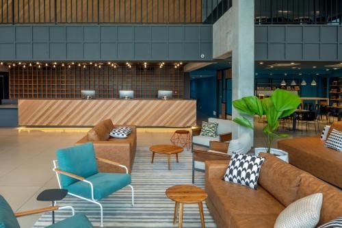 Sala de estar moderna do hotel com assentos em tons de azul-petróleo e bege, mesas de madeira e um grande vaso de planta.