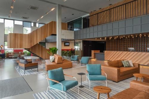 Hall de entrada moderno com cadeiras verde-azuladas, sofás bege, detalhes em madeira e grandes janelas em um interior amplo e iluminado.