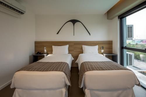 Quarto de hotel moderno com duas camas de solteiro, cabeceira de madeira e uma grande janela com vista para a cidade.