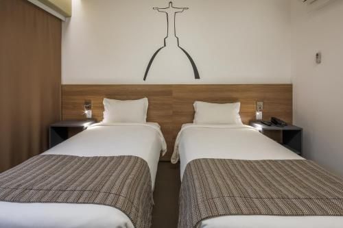 Duas camas de solteiro em um quarto de hotel simples, com lençóis brancos, cabeceira de madeira e um cabideiro de parede acima.