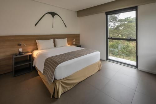 Quarto moderno com cama grande, cabeceira de madeira, mesa de cabeceira e janela alta com vista para as árvores.