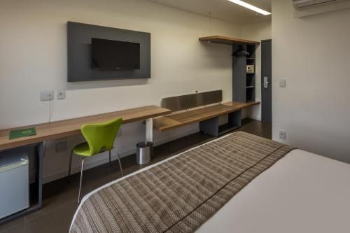 Quarto moderno com TV de parede, escrivaninha comprida, cadeira verde e cama em primeiro plano.