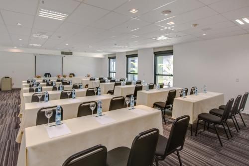 Sala de conferências vazia com fileiras de mesas bege e cadeiras pretas sob luzes fortes no teto.