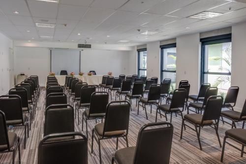 Sala de conferências vazia com fileiras de cadeiras e mesas pretas perto de grandes janelas.