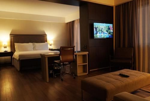 Quarto de hotel com cama grande, escrivaninha, TV de parede e cortinas escuras em ambiente com iluminação aconchegante.