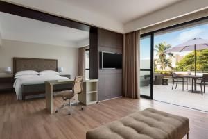 Quarto de hotel moderno com cama, escrivaninha, TV e varanda com vista para palmeiras e um pátio externo.