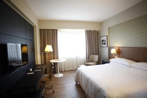 Quarto de hotel moderno com cama, TV, luminária e janela com cortinas translúcidas.