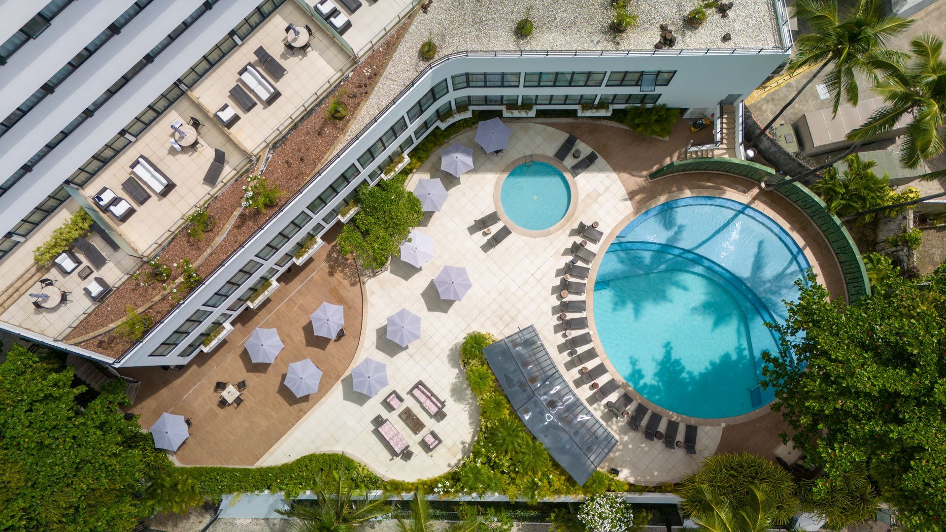 Piscina externa do hotel ao lado de um prédio branco moderno, com espreguiçadeiras e guarda-sóis.