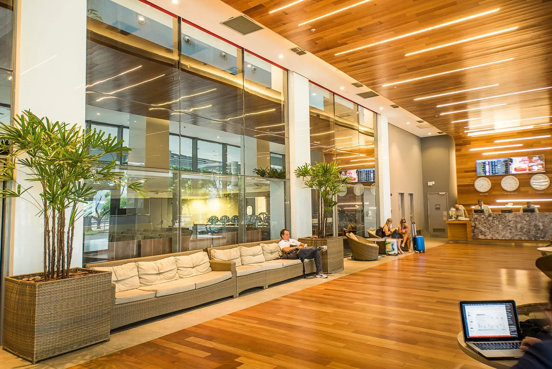 Sala de espera moderna de aeroporto com assentos, piso de madeira, paredes de vidro e balcão de check-in ao fundo.