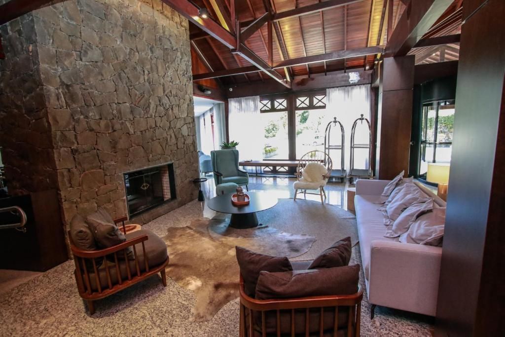 Sala de estar aconchegante com lareira de pedra, teto de madeira, sofá branco e grandes janelas com vista para a vegetação.