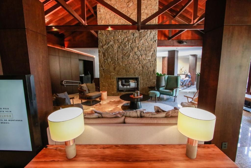 Sala de estar da pousada com lareira de pedra, vigas de madeira e abajures aconchegantes emoldurando a vista.