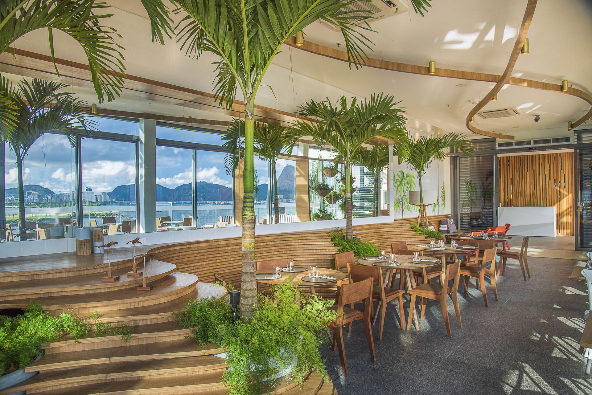 Interior luminoso de restaurante tropical com mesas de madeira, palmeiras em vasos e janelas com vista para o mar.