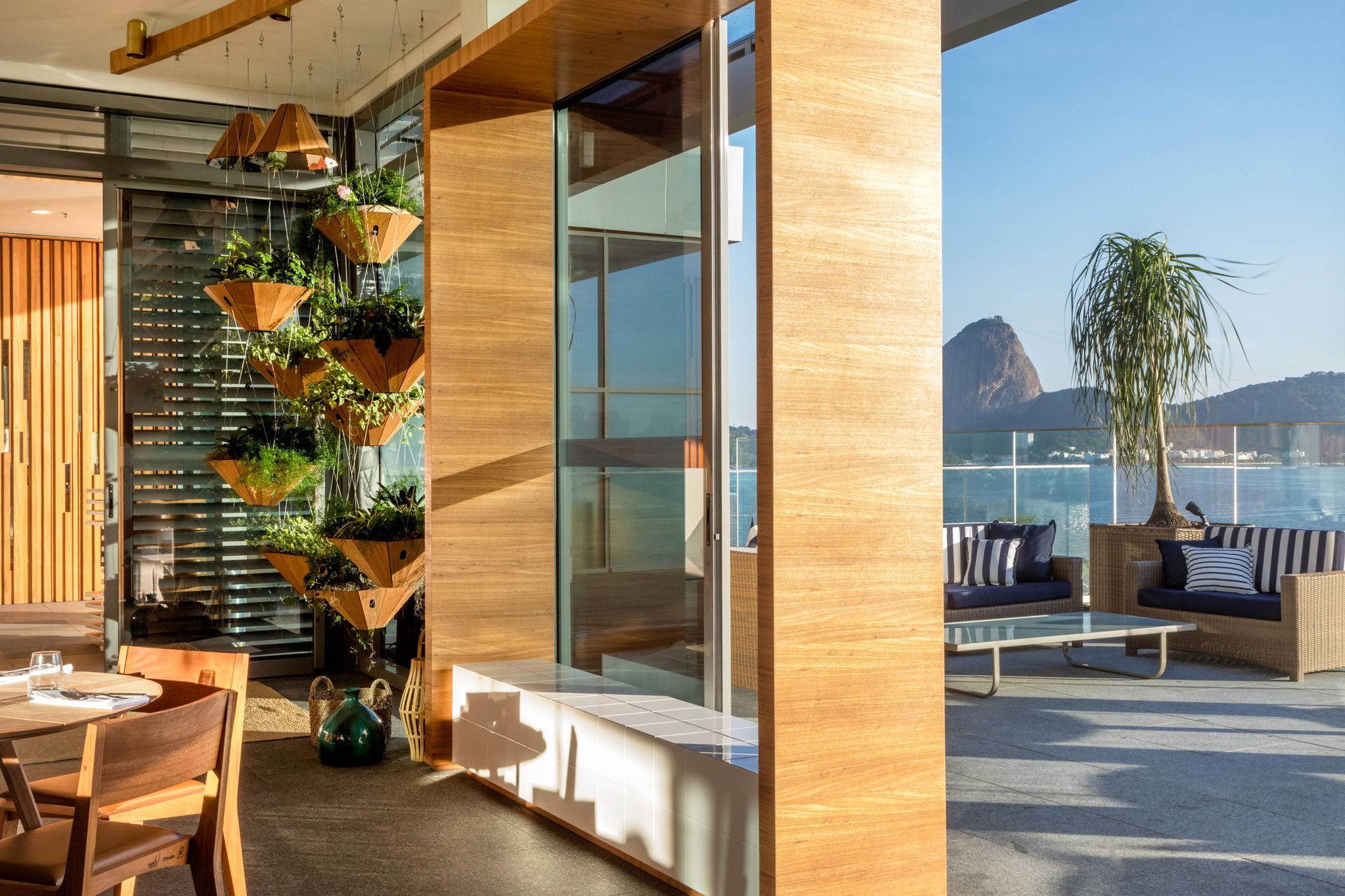 Lounge moderno com varanda, vista para o mar, plantas suspensas e decoração em madeira aconchegante.