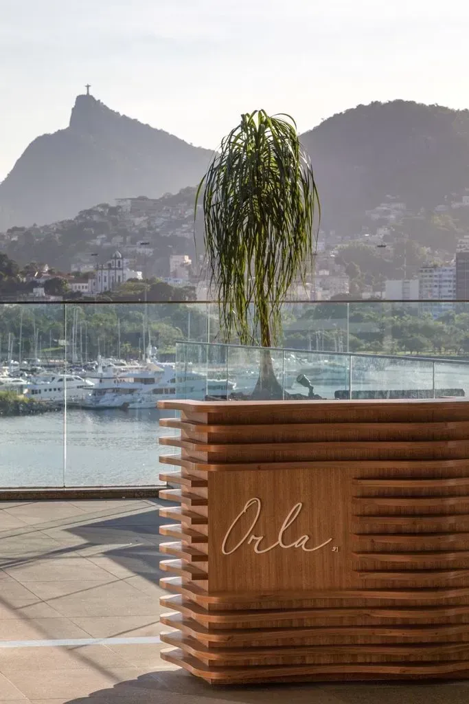 Mesa de madeira ao ar livre com planta suspensa, com vista para o porto e as montanhas ao pôr do sol.
