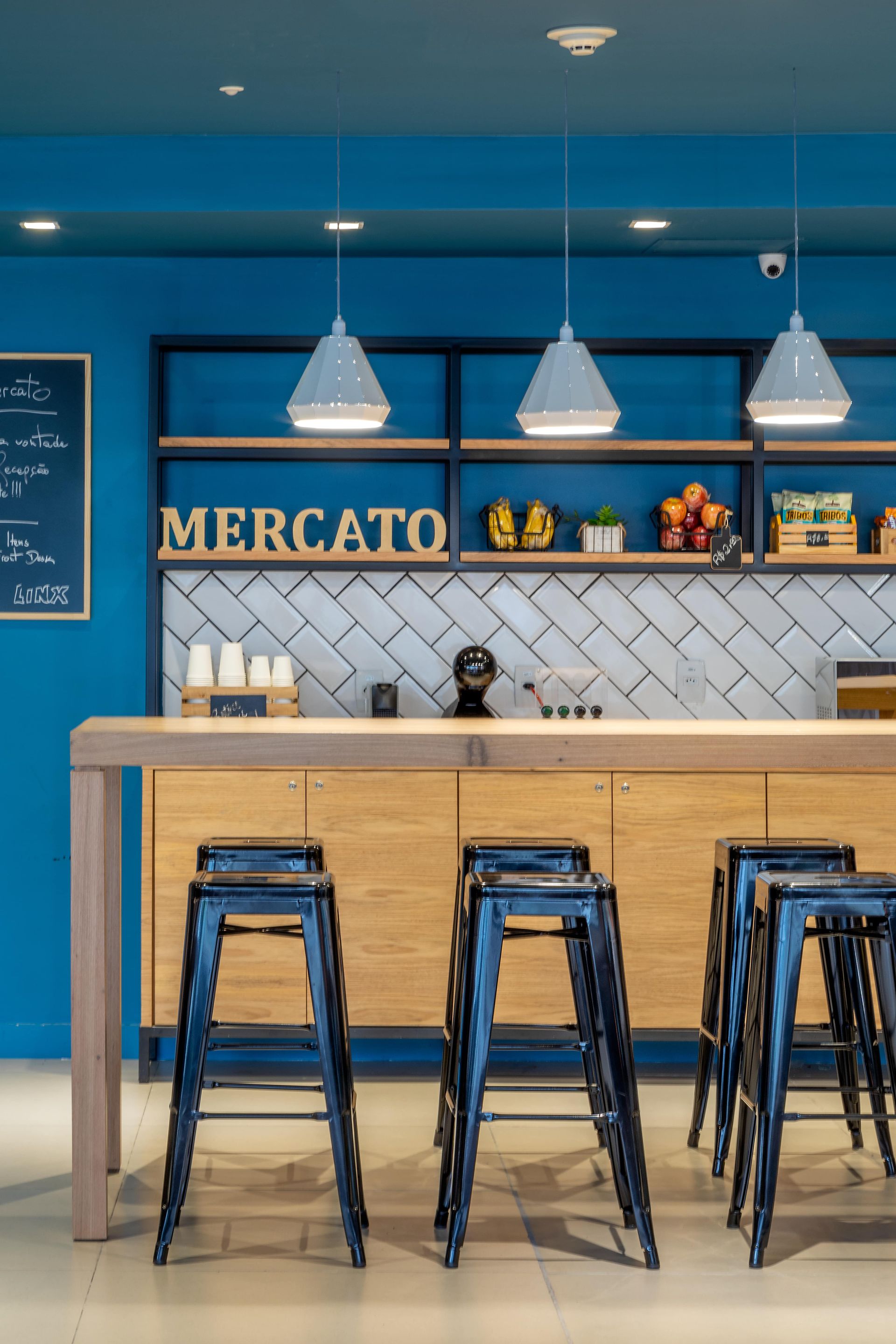 Interior azul de um café com balcão de madeira, banquetas de metal, luminárias pendentes e uma placa com a palavra “MERCATO” atrás do balcão.