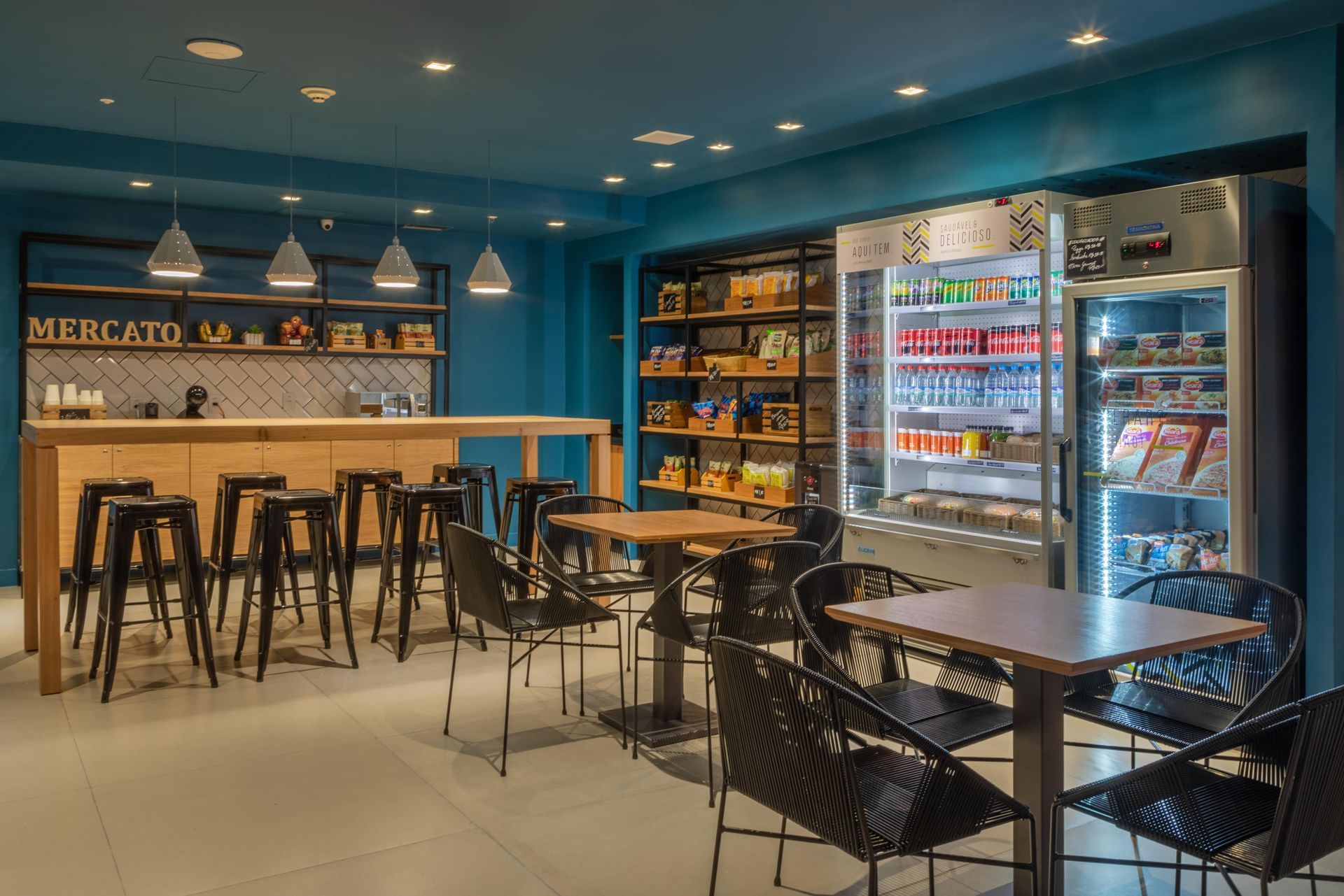 Interior de café moderno com paredes azuis, bancos de bar, mesas e um expositor refrigerado de bebidas.