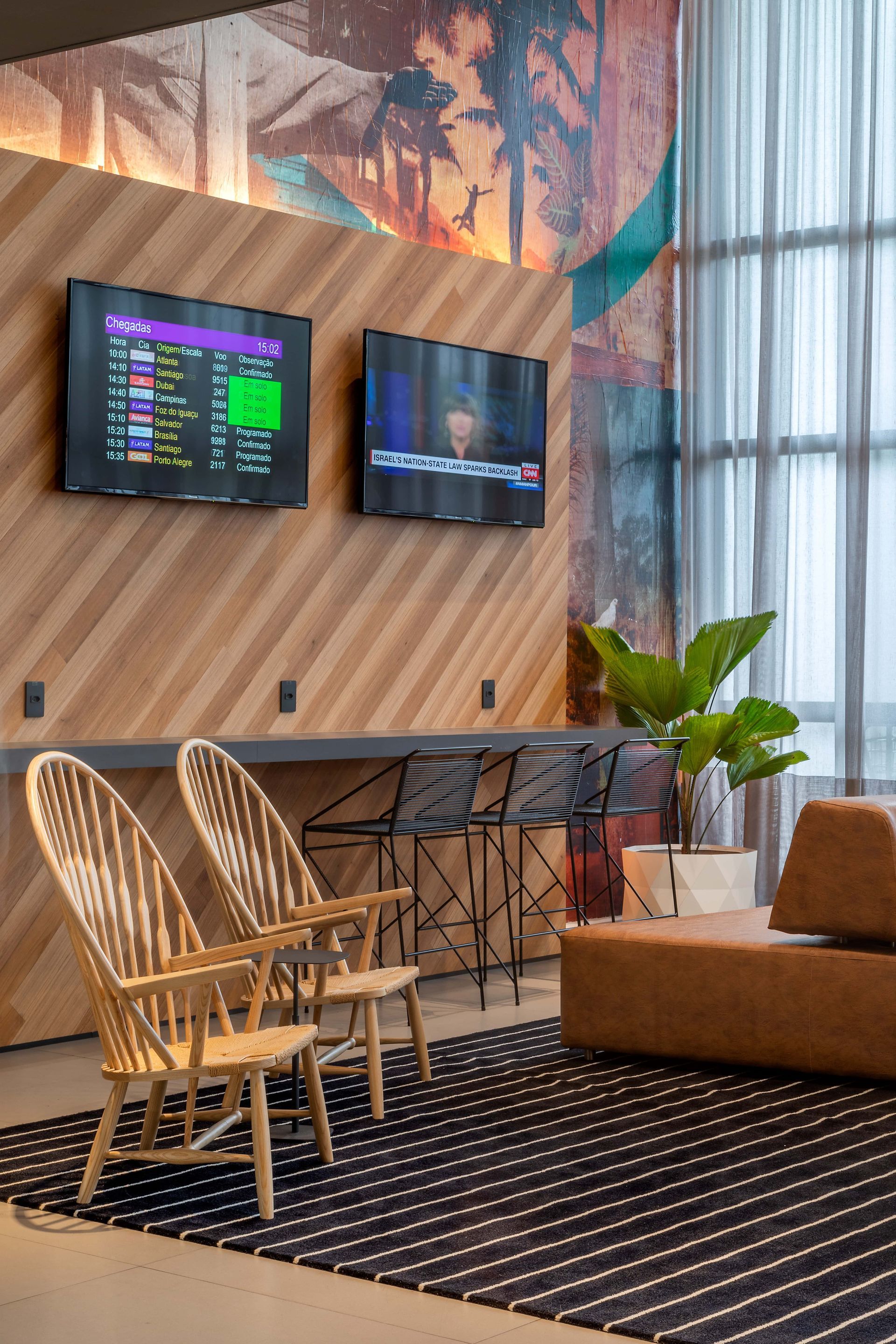 Sala de estar moderna com cadeiras de vime, TVs fixadas na parede, parede de madeira, plantas e um sofá junto à janela.